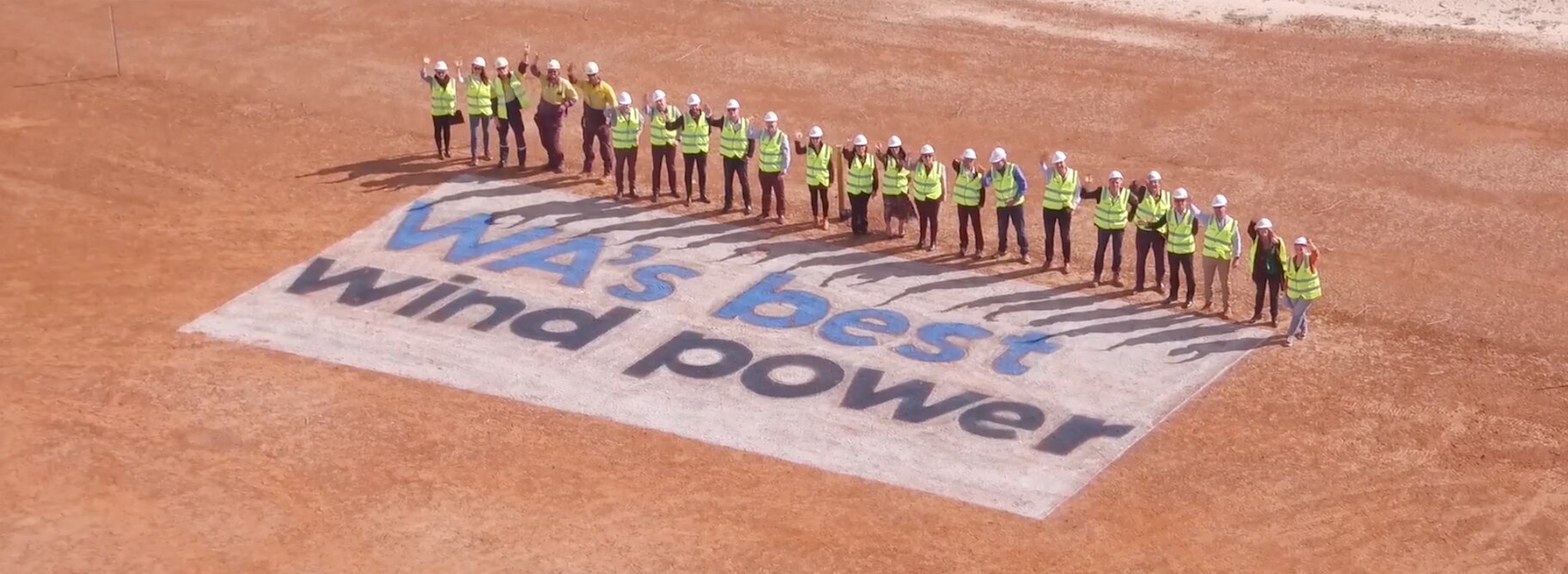An aerial shot of several workers standing in a line wearing hi-vis and hard hats looking up and waving at the camera. The ground is dry and brown-orange in colour. In front of them is a painted sign in a white rectangle reading 'WA's best wind power'